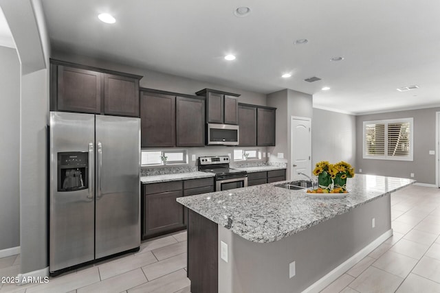 kitchen with appliances with stainless steel finishes, sink, light stone counters, dark brown cabinetry, and a center island with sink