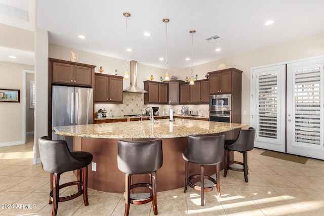 kitchen with stainless steel appliances, wall chimney range hood, hanging light fixtures, an island with sink, and a breakfast bar area