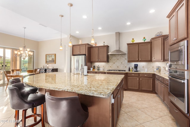 kitchen with stainless steel appliances, hanging light fixtures, light tile patterned floors, a kitchen island with sink, and wall chimney range hood