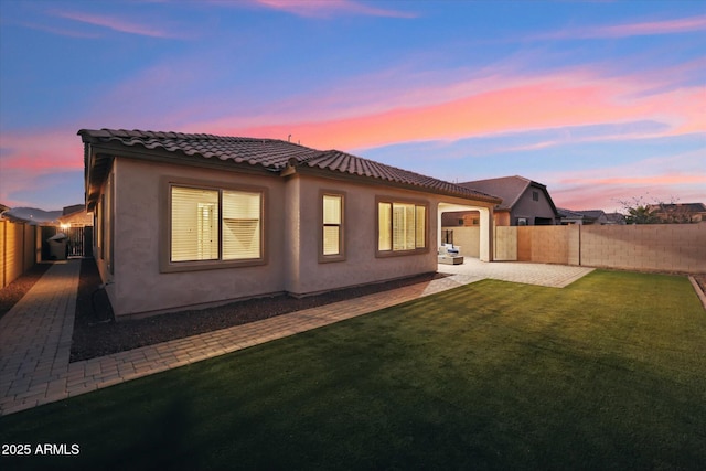 back of house at dusk featuring a fenced backyard, a tile roof, a lawn, stucco siding, and a patio area