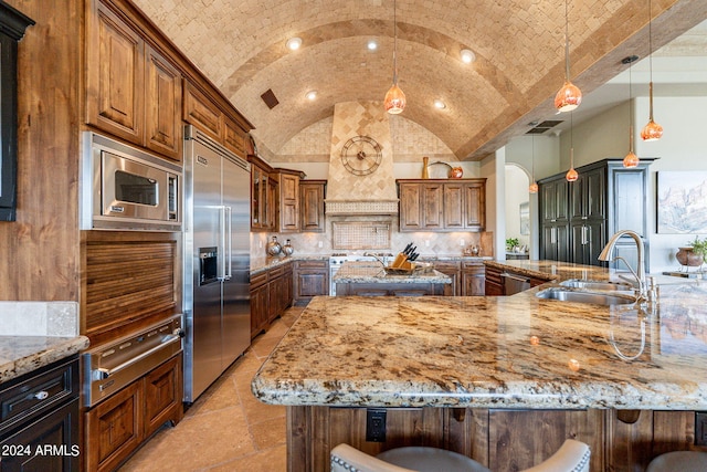 kitchen with light stone counters, pendant lighting, built in appliances, a kitchen breakfast bar, and vaulted ceiling