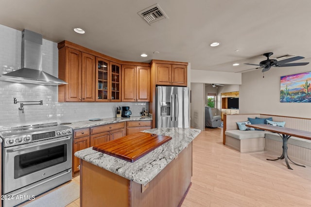 kitchen with ceiling fan, stainless steel appliances, wall chimney range hood, light stone counters, and a kitchen island