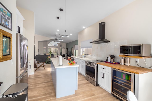 kitchen with wall chimney range hood, wine cooler, an island with sink, appliances with stainless steel finishes, and decorative light fixtures