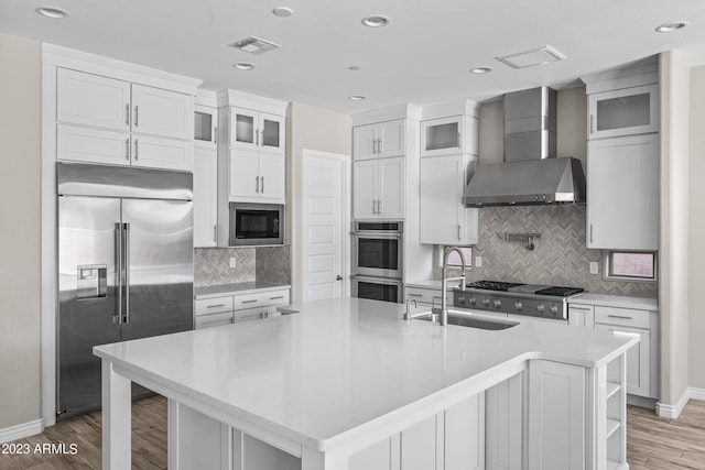 kitchen featuring light wood-type flooring, wall chimney exhaust hood, a kitchen island with sink, built in appliances, and white cabinetry