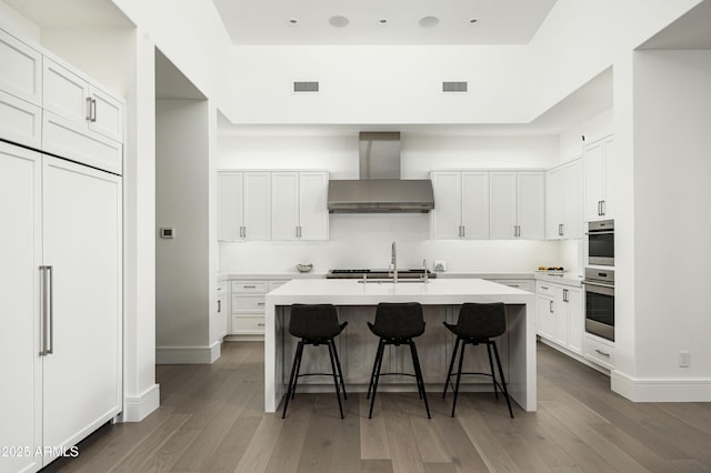 kitchen with white cabinetry, a center island with sink, wall chimney exhaust hood, and a kitchen bar