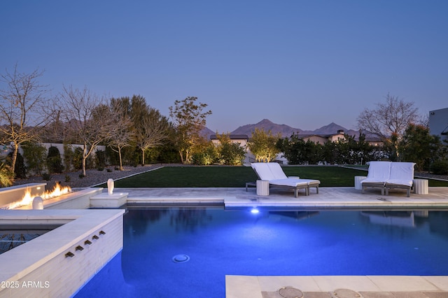 pool at dusk with a patio, a mountain view, a fire pit, and a lawn