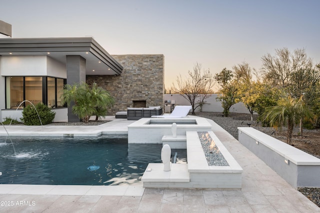 pool at dusk featuring a patio area, pool water feature, and an outdoor living space with a fireplace