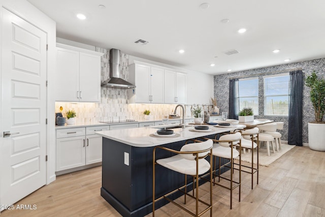 kitchen featuring wall chimney range hood, a kitchen island with sink, visible vents, and white cabinets