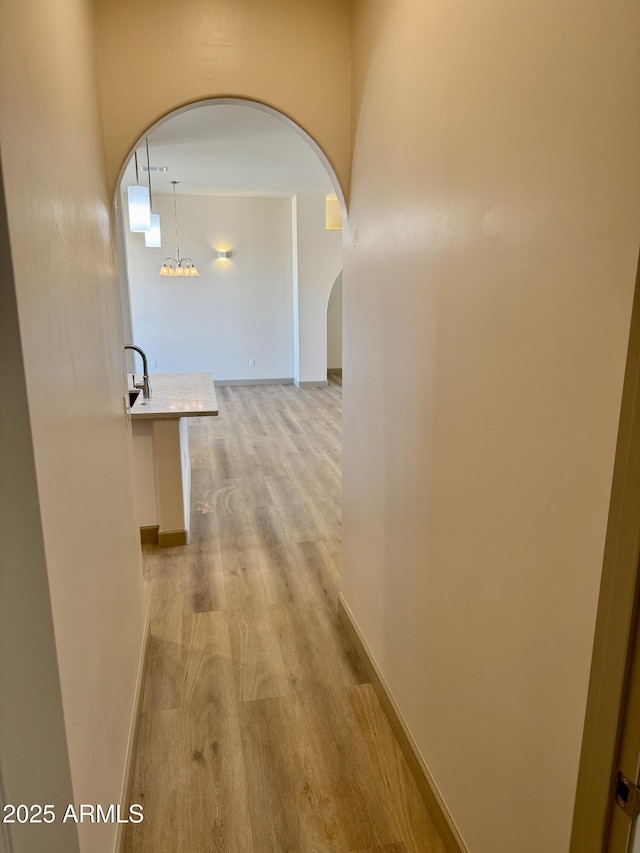 corridor with arched walkways, light wood finished floors, an inviting chandelier, a sink, and baseboards