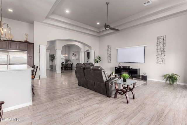 living room with a tray ceiling, decorative columns, light hardwood / wood-style flooring, ceiling fan with notable chandelier, and a towering ceiling