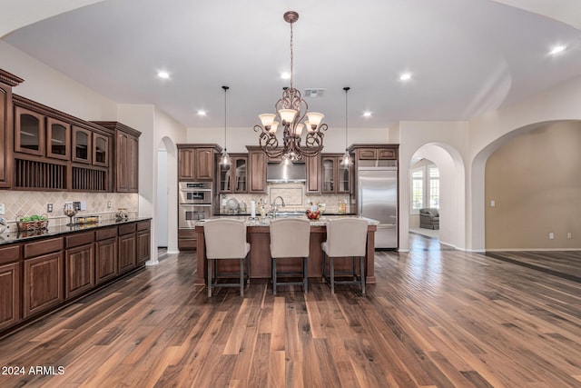 kitchen with dark hardwood / wood-style flooring, pendant lighting, decorative backsplash, a center island with sink, and appliances with stainless steel finishes