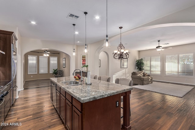 kitchen with a kitchen island with sink, dark wood-type flooring, sink, hanging light fixtures, and light stone countertops