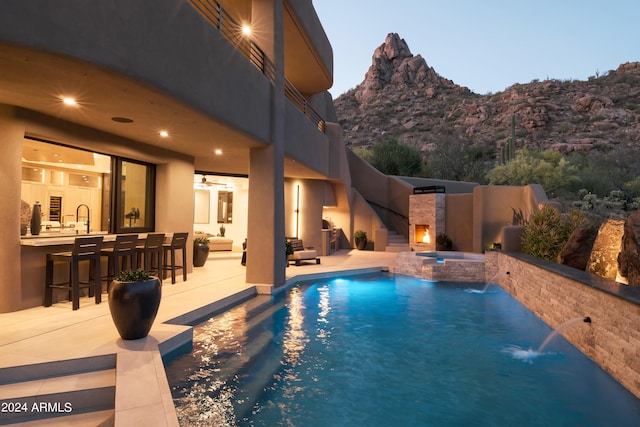 pool at dusk featuring an outdoor stone fireplace, pool water feature, a mountain view, a patio, and an outdoor bar