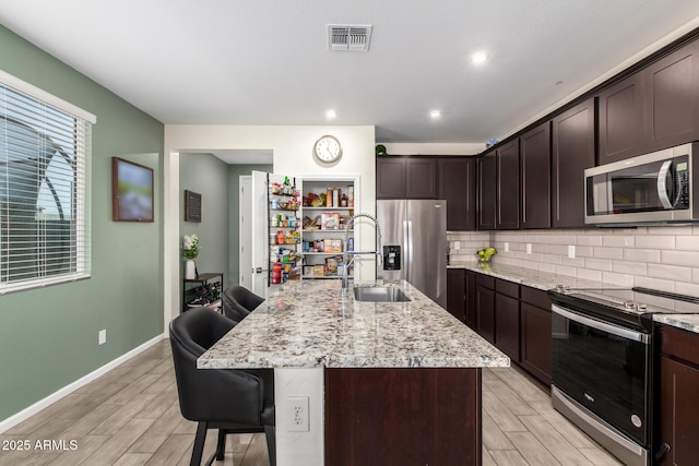 kitchen featuring dark brown cabinets, backsplash, light stone countertops, appliances with stainless steel finishes, and a kitchen breakfast bar