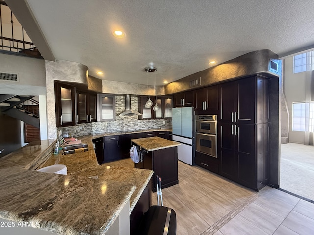 kitchen featuring pendant lighting, glass insert cabinets, a sink, wall chimney range hood, and a peninsula