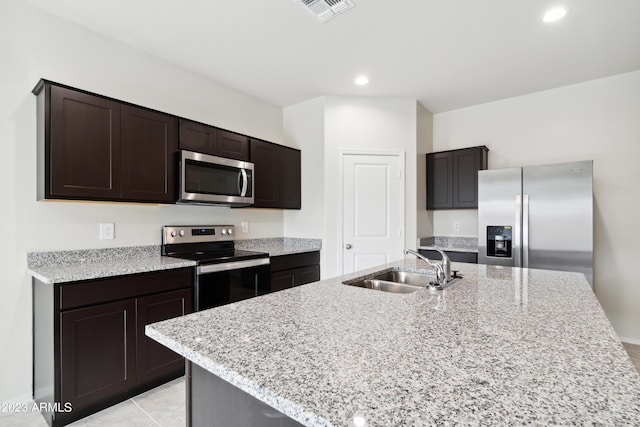 kitchen with stainless steel appliances, a kitchen island with sink, dark brown cabinets, light stone countertops, and sink