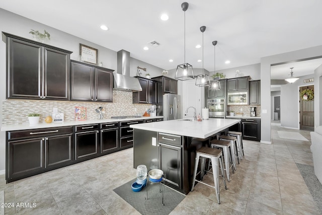 kitchen featuring hanging light fixtures, wall chimney exhaust hood, an island with sink, a breakfast bar area, and stainless steel appliances