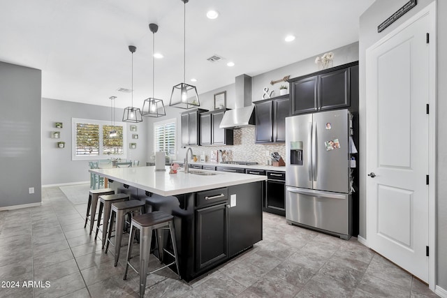 kitchen with a kitchen island with sink, wall chimney range hood, hanging light fixtures, sink, and appliances with stainless steel finishes