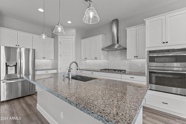 kitchen with stainless steel appliances, wall chimney range hood, a center island with sink, white cabinets, and hanging light fixtures