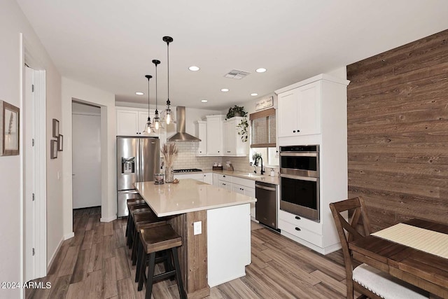 kitchen with wall chimney exhaust hood, stainless steel appliances, decorative light fixtures, hardwood / wood-style flooring, and a kitchen island