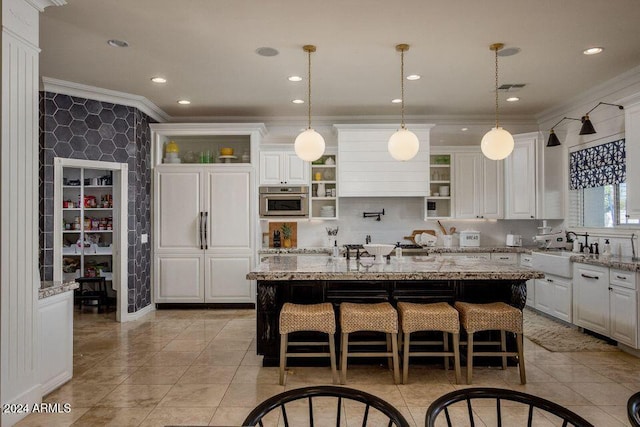 kitchen with white cabinets, light stone counters, ornamental molding, and an island with sink