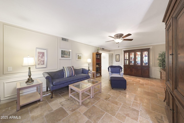 living area featuring ceiling fan, visible vents, crown molding, stone tile flooring, and a decorative wall