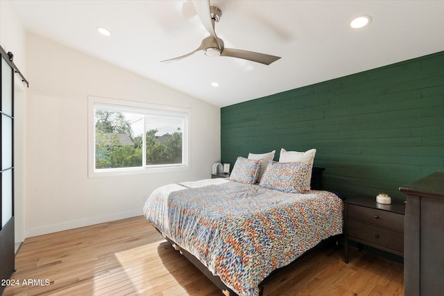 bedroom with vaulted ceiling, light wood-type flooring, wooden walls, ceiling fan, and a barn door