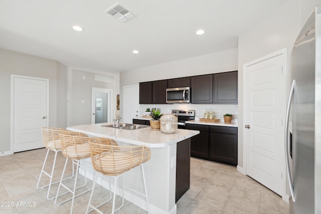 kitchen featuring sink, a kitchen island with sink, dark brown cabinets, stainless steel appliances, and light stone countertops