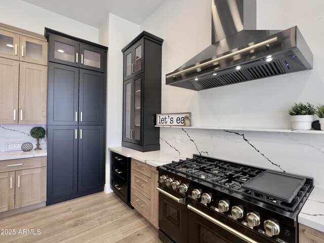kitchen featuring wall chimney range hood, range with two ovens, decorative backsplash, light wood finished floors, and light stone counters