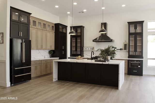 kitchen with black appliances, wall chimney range hood, light countertops, light wood-style flooring, and decorative backsplash