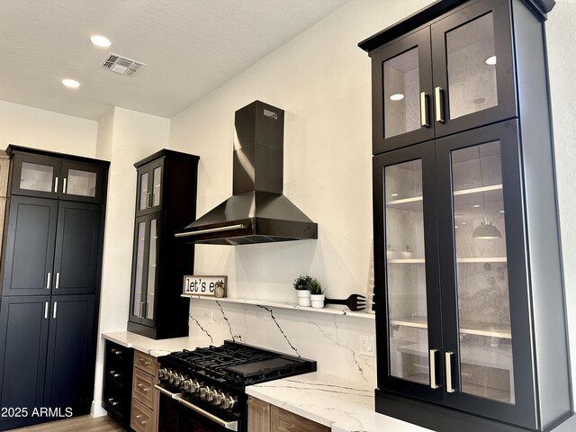 kitchen featuring black range with gas cooktop, wall chimney range hood, light stone counters, tasteful backsplash, and glass insert cabinets