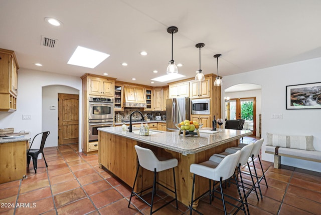 kitchen featuring a large island with sink, tasteful backsplash, decorative light fixtures, light stone counters, and stainless steel appliances