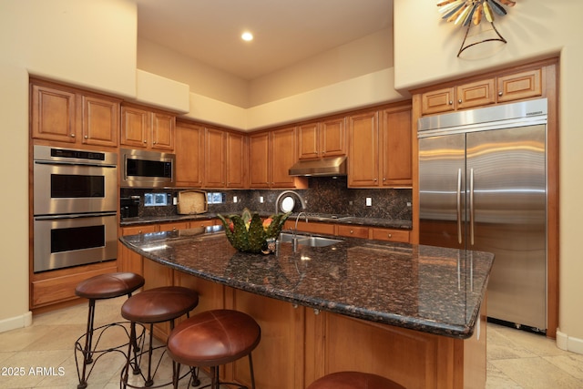 kitchen featuring brown cabinets, decorative backsplash, built in appliances, dark stone counters, and under cabinet range hood