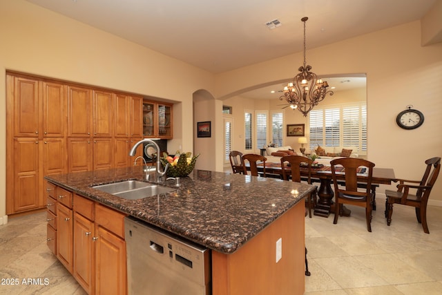 kitchen featuring arched walkways, brown cabinetry, an island with sink, stainless steel dishwasher, and a sink