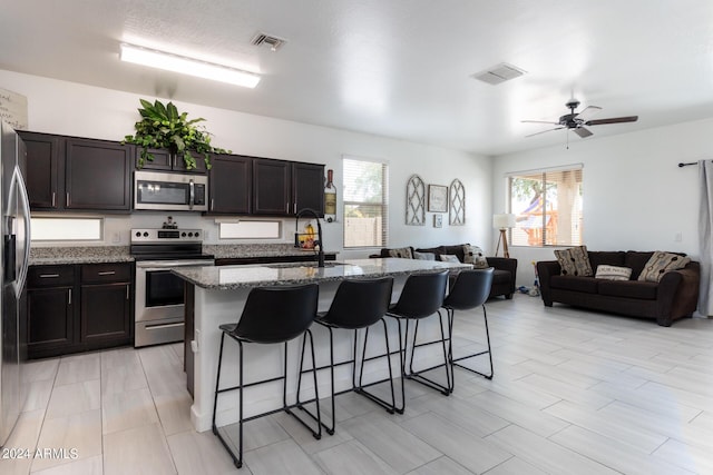 kitchen featuring appliances with stainless steel finishes, sink, a kitchen island with sink, dark brown cabinetry, and light stone countertops