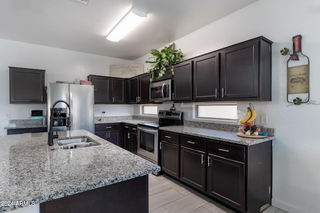 kitchen with sink, dark brown cabinets, stainless steel appliances, light stone countertops, and a center island with sink