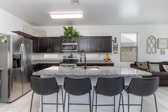 kitchen featuring dark brown cabinetry, sink, light stone counters, an island with sink, and stainless steel appliances