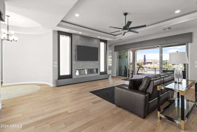 living room featuring light wood-type flooring, a tray ceiling, a large fireplace, and ceiling fan with notable chandelier