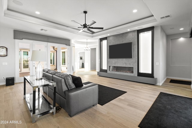 living room featuring light hardwood / wood-style floors, ornamental molding, ceiling fan with notable chandelier, a large fireplace, and a tray ceiling