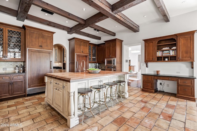 kitchen featuring beam ceiling, butcher block counters, a kitchen breakfast bar, built in appliances, and a center island with sink