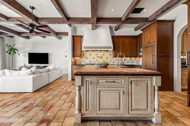 kitchen with premium range hood, a kitchen island with sink, beamed ceiling, decorative backsplash, and butcher block countertops