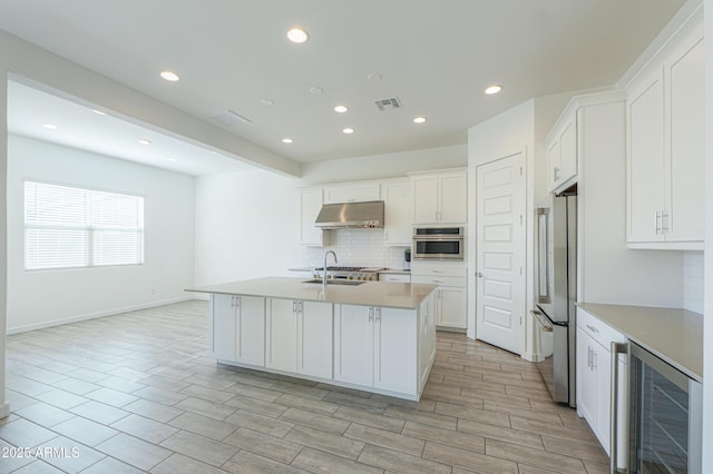 kitchen featuring wine cooler, decorative backsplash, a center island with sink, white cabinets, and appliances with stainless steel finishes