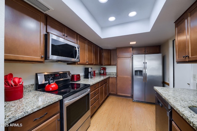 kitchen with stainless steel appliances, a raised ceiling, light stone counters, and light hardwood / wood-style flooring