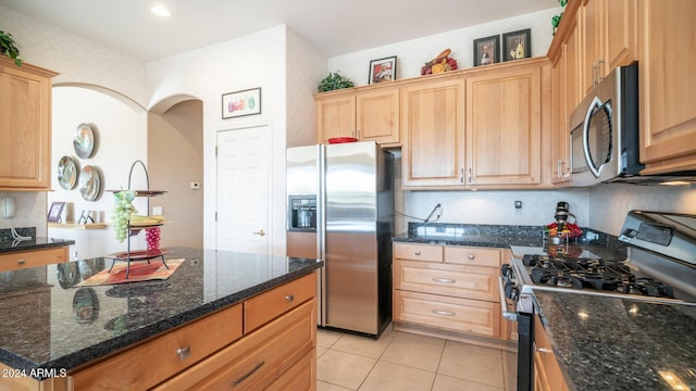 kitchen with light brown cabinetry, stainless steel appliances, light tile patterned floors, dark stone countertops, and a center island