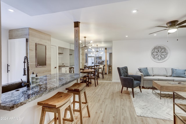 kitchen featuring a breakfast bar, light wood-type flooring, dark stone countertops, ceiling fan with notable chandelier, and pendant lighting