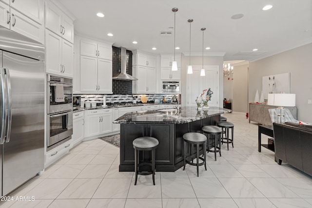 kitchen featuring wall chimney exhaust hood, dark stone countertops, pendant lighting, a kitchen island with sink, and appliances with stainless steel finishes