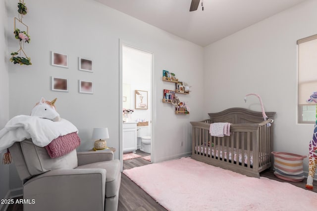 bedroom featuring wood finished floors, a crib, ensuite bathroom, and a ceiling fan