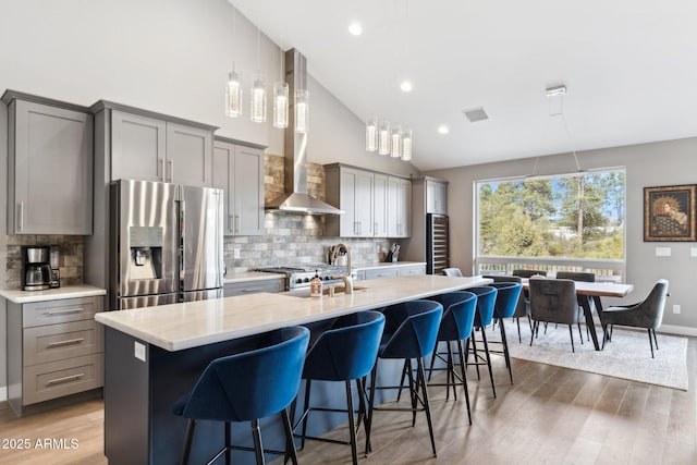 kitchen with stainless steel refrigerator with ice dispenser, a kitchen bar, a center island with sink, gray cabinets, and wall chimney range hood