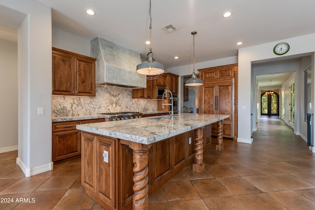 kitchen with a kitchen island with sink, hanging light fixtures, custom range hood, backsplash, and light stone countertops