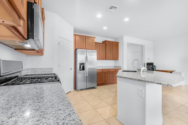 kitchen with stove, sink, stainless steel fridge, light stone countertops, and an island with sink